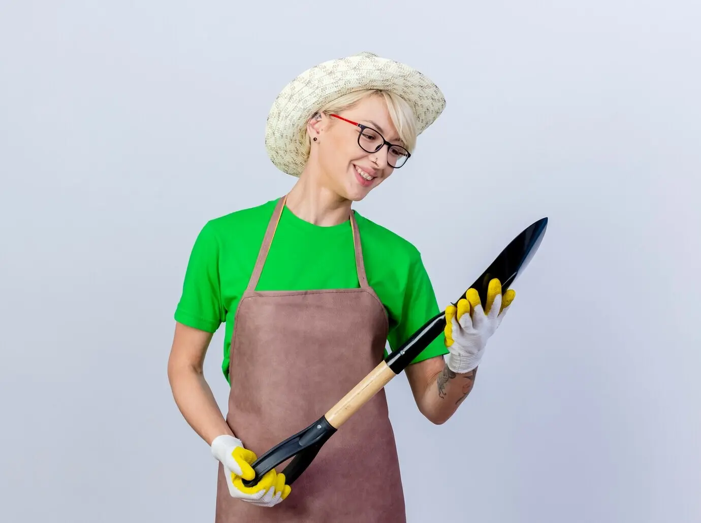 Junge Gärtnerin mit kurzen Haaren, in Schürze und Hut, hält eine Schaufel, schaut sie an und lächelt glücklich.