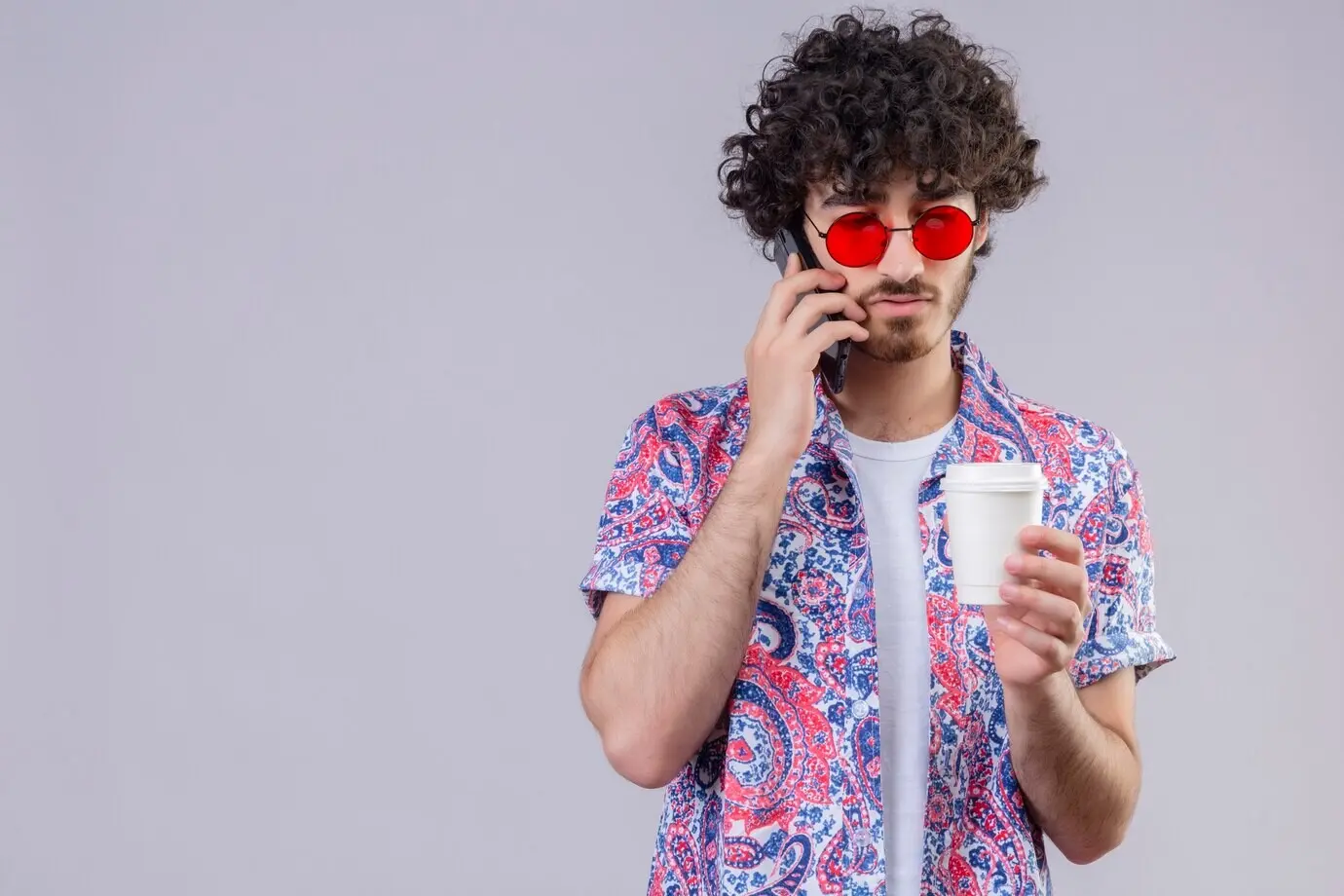 Junger gutaussehender lockiger Reisender trägt eine Sonnenbrille, spricht am Telefon, hält einen Plastik-Kaffeebecher und schaut ihn an, vor einer isolierten weißen Wand mit Copyspace.