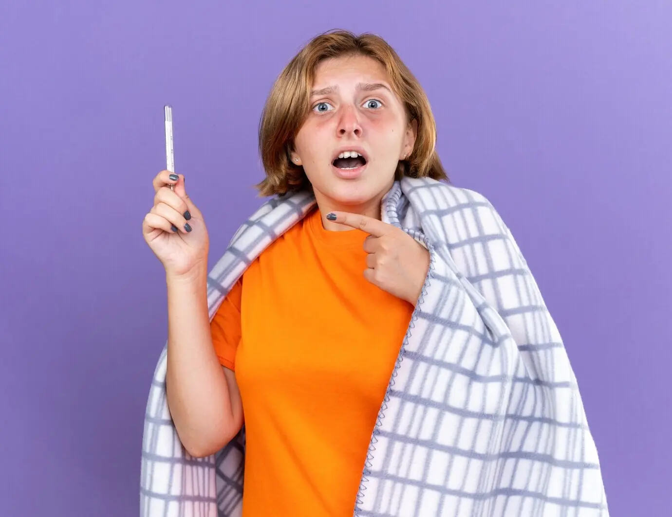 Eine gesundheitlich angeschlagene junge Frau, in eine warme Decke gehüllt, fühlt sich krank, leidet an Grippe und hat Fieber; sie misst ihre Temperatur mit einem Thermometer, zeigt darauf und wirkt besorgt, während sie vor einer violetten Wand steht.