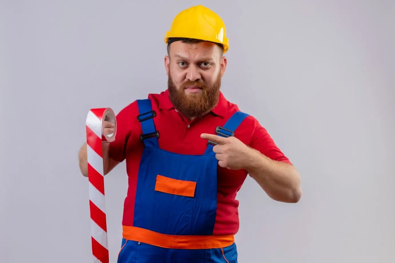 Junger, bärtiger Bauarbeiter in Arbeitskleidung und Sicherheitshelm hält Klebeband, zeigt mit dem Finger darauf und schaut unzufrieden.