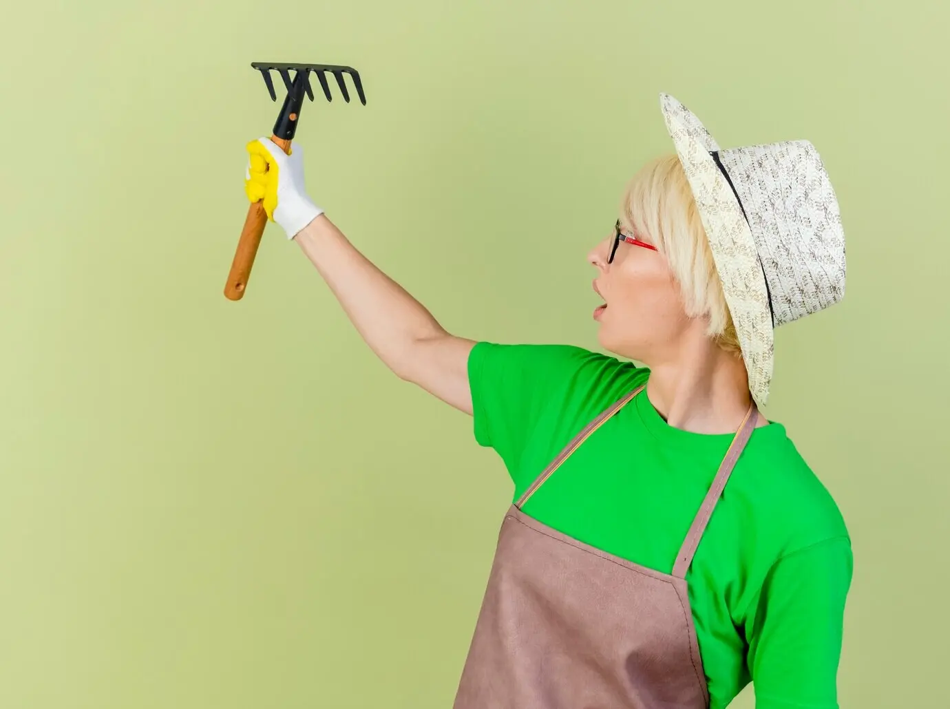 Eine junge Gärtnerin mit kurzen Haaren trägt Schürze, Hut und Gummihandschuhe, hält eine Miniharke und schaut sie überrascht an, während sie vor hellem Hintergrund steht.