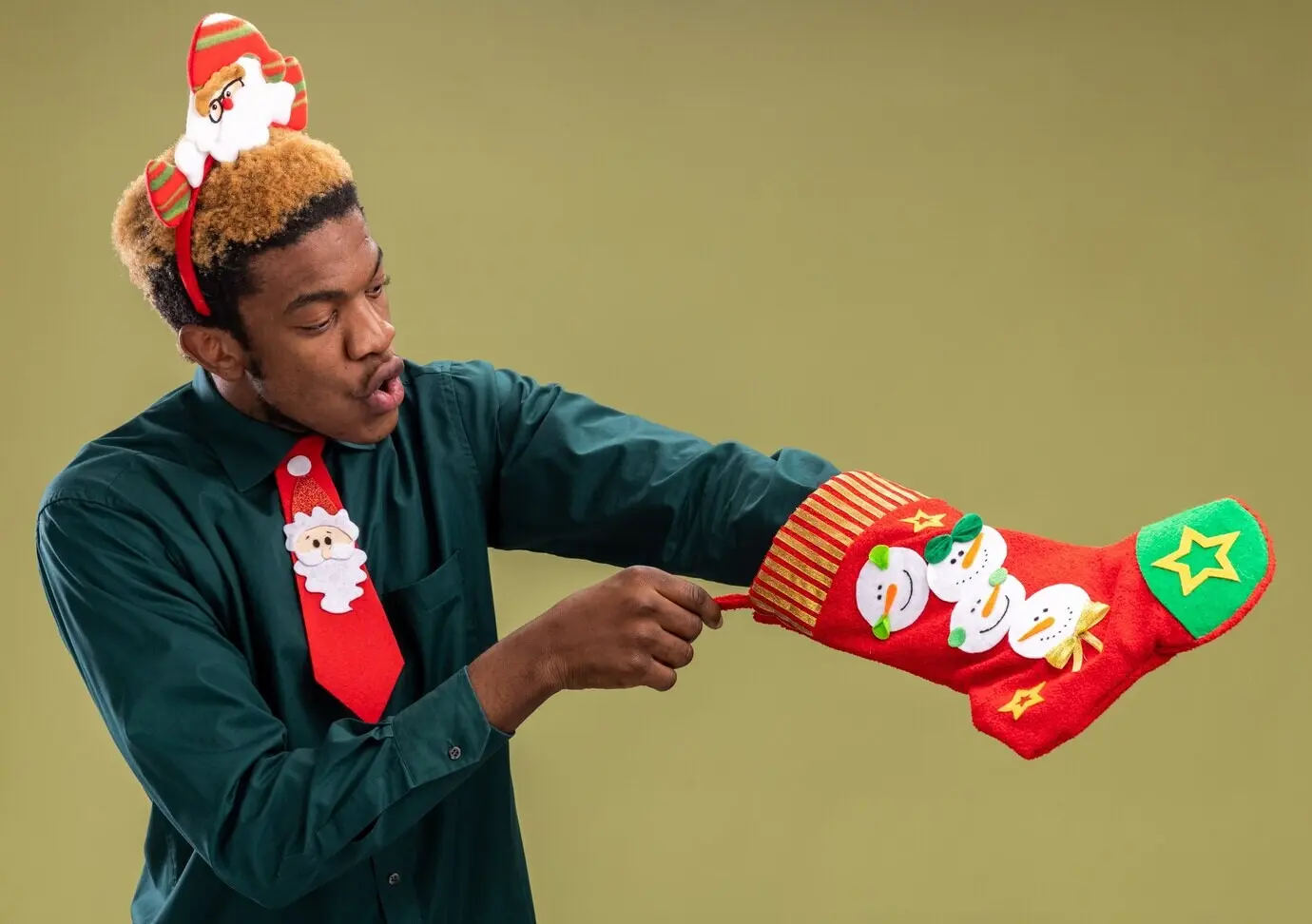 Afroamerikanischer Mann mit lustigem Weihnachtsmann-Accessoire und roter Krawatte hält einen Weihnachtsstrumpf, den er überrascht betrachtet, und steht vor einer grünen Wand.