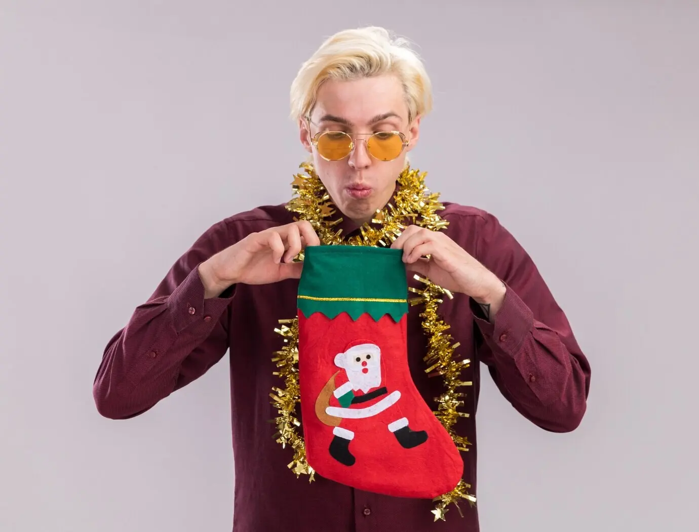Neugieriger junger blonder Mann mit Brille und Lametta-Girlande um den Hals hält einen Weihnachtsstrumpf und blickt hinein, isoliert vor weißem Hintergrund.