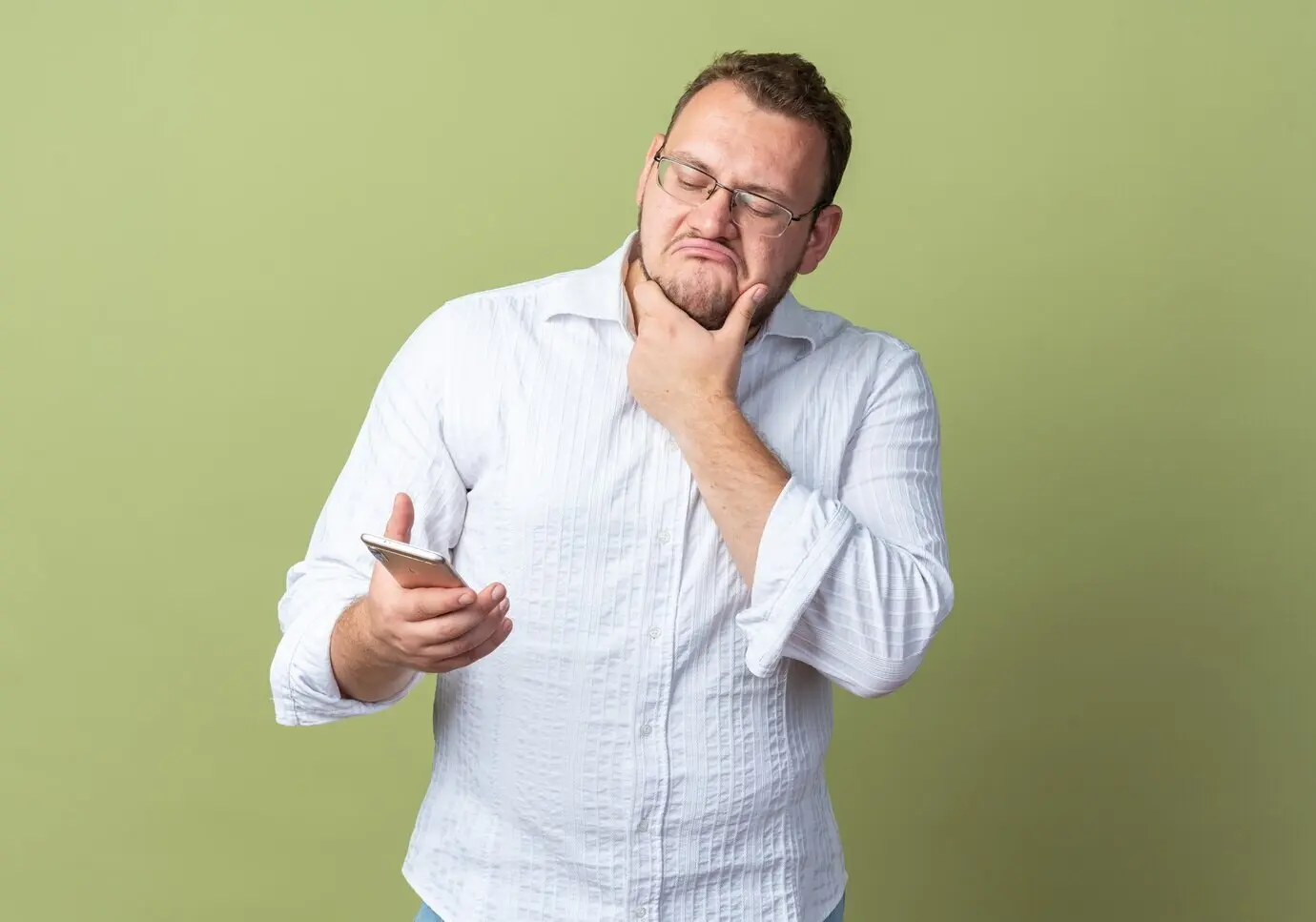 Ein Mann im weißen Hemd mit Brille hält ein Smartphone und schaut mit skeptischem Ausdruck darauf, während er vor einer grünen Wand steht.